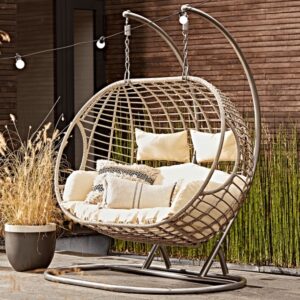 Outdoor egg-shaped wicker hanging chair with cushions on a patio, featuring a cozy beige design and round metal base
